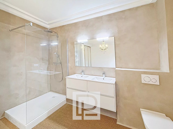 Modern bathroom with glass-walled shower, white double sink vanity, and a large mirror reflecting a chandelier beyond the room.