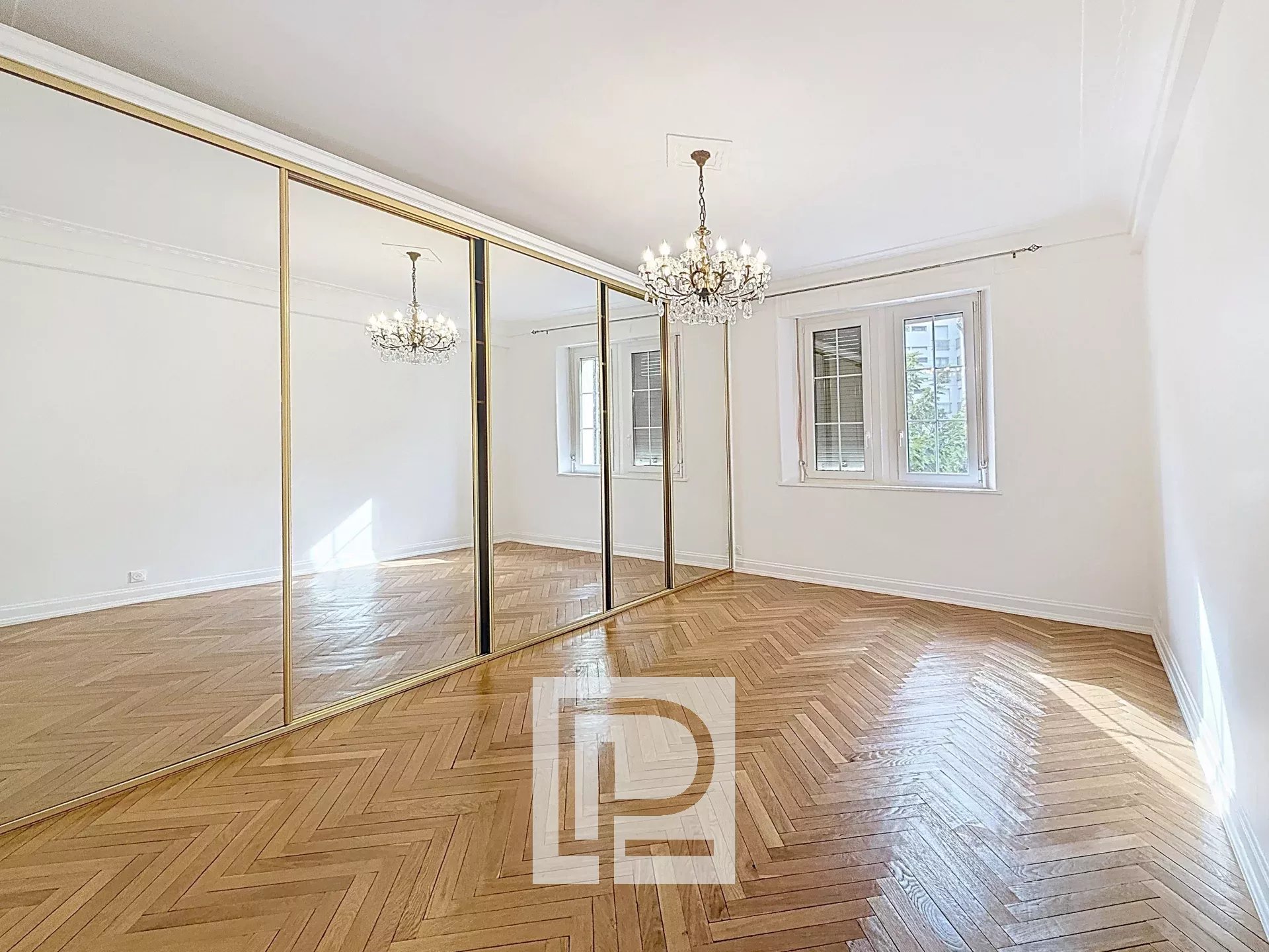 Bright empty room with herringbone wood floor, gold-framed mirrored closets, and a crystal chandelier.