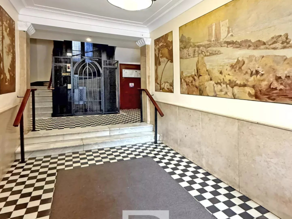 Hotel lobby hallway with a black-and-white checkered floor, a brown runner rug, and wall paintings; an ornate ceiling light hangs above and an iron elevator gate is at the far end.