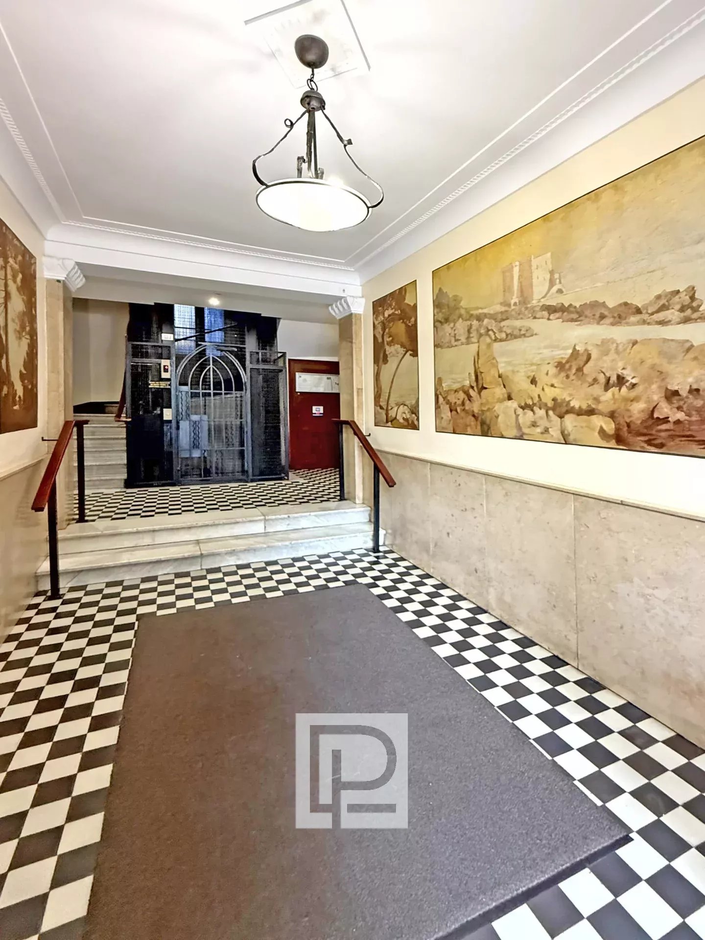 Hotel lobby hallway with a black-and-white checkered floor, a brown runner rug, and wall paintings; an ornate ceiling light hangs above and an iron elevator gate is at the far end.