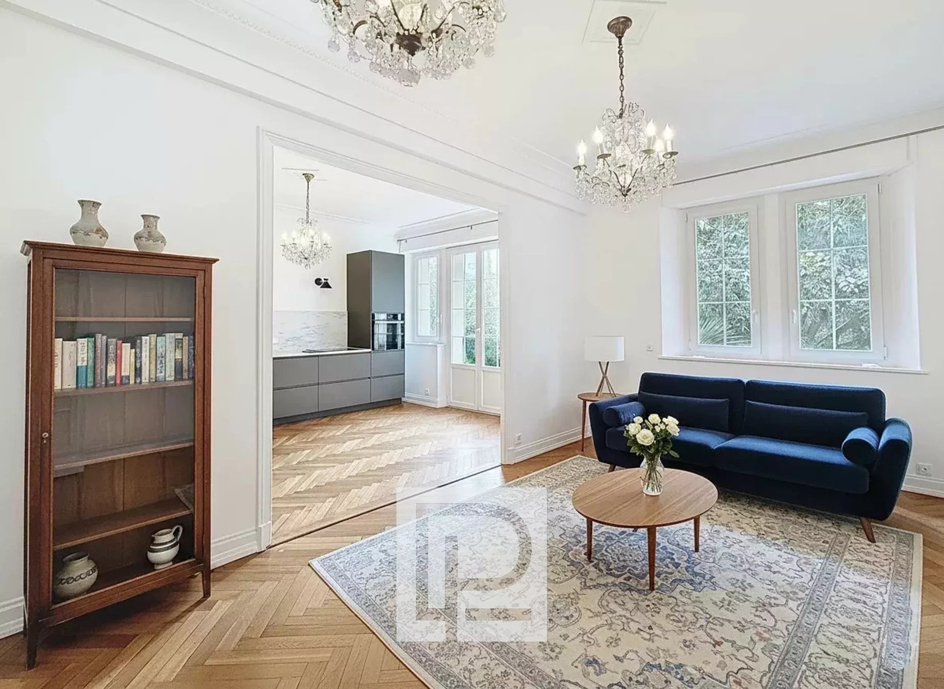 Bright living room with a royal blue sofa, round wooden coffee table, and a patterned rug. Large windows let in natural light; candlestick overhead.