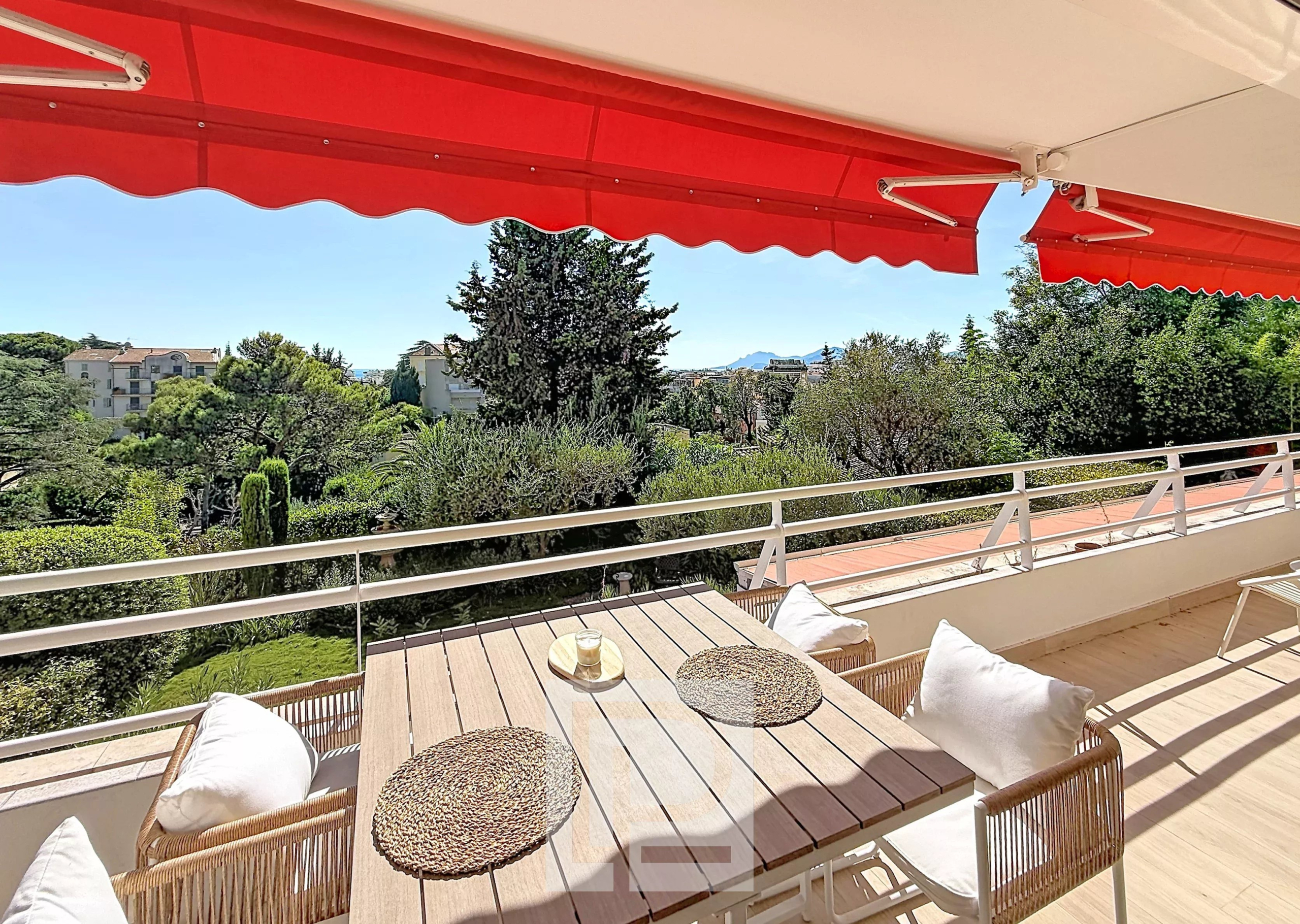 Terrasse d’un appartement à Cannes Terrasse d'un appartement rénové avec vue sur la verdure à Cannes, mobilier en bois et parasols rouges.