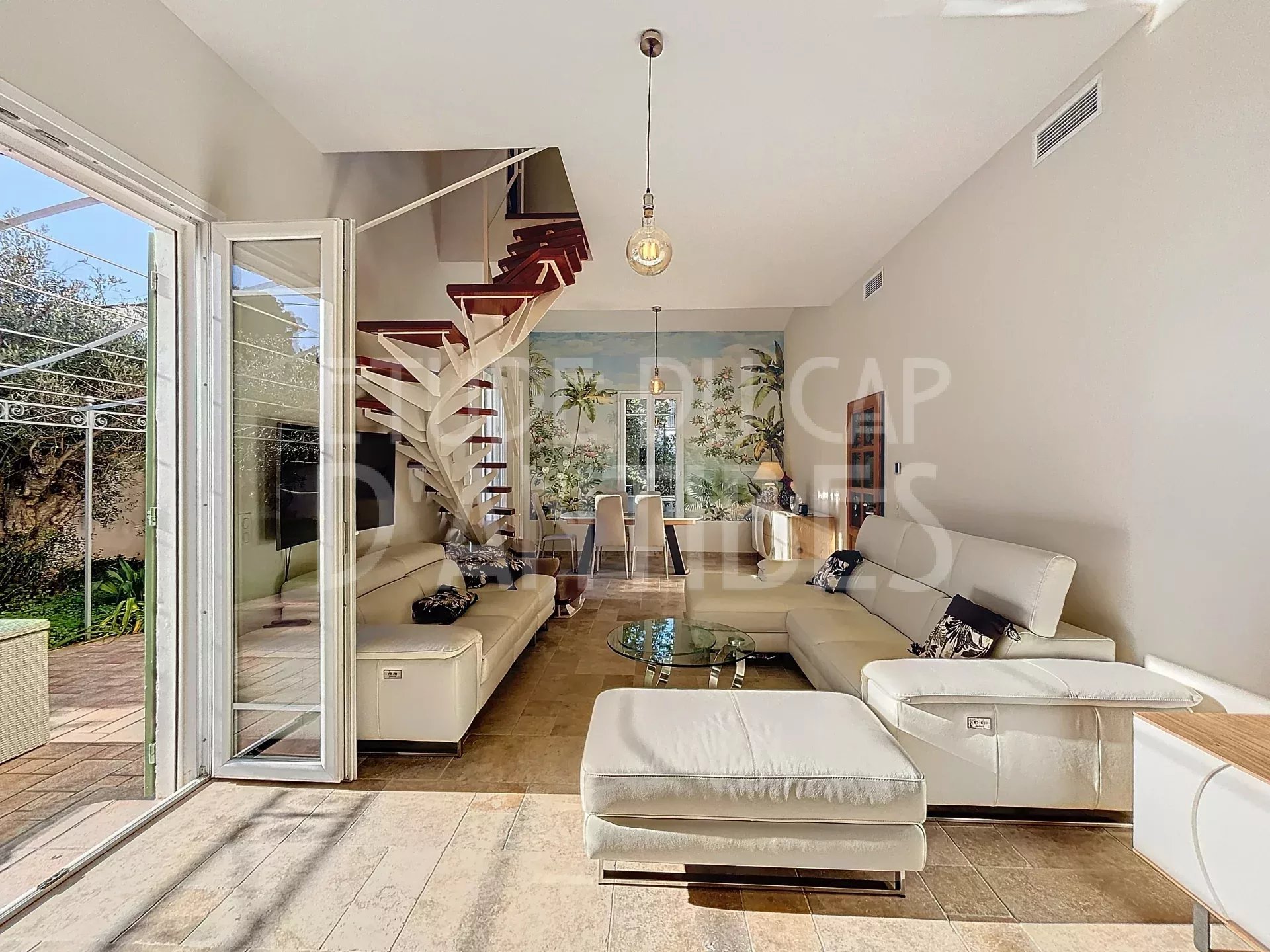 Bright open-plan living room with white sectional sofas, glass coffee table, and a spiral staircase; tropical mural on back wall beside dining area.