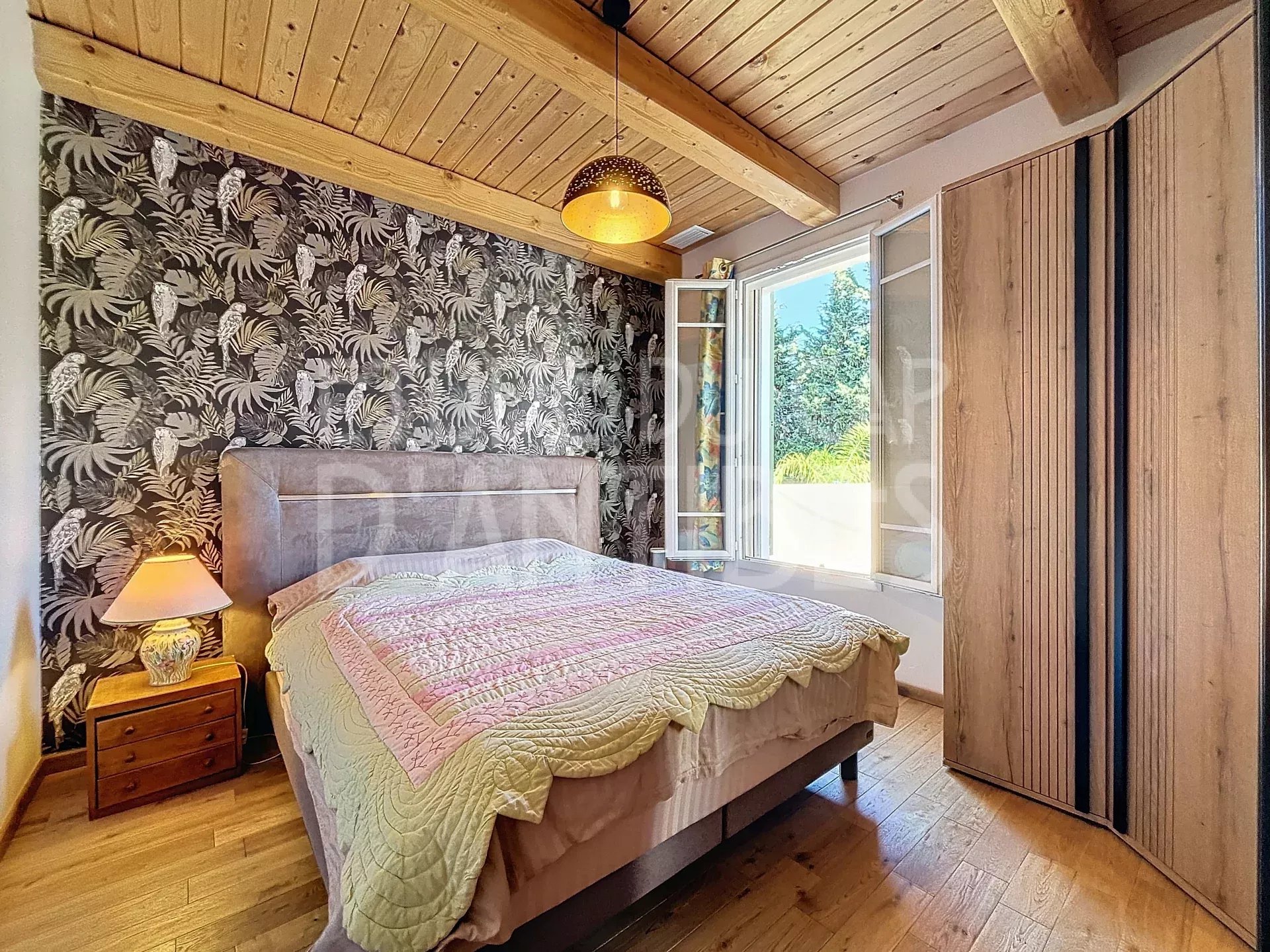 Bedroom with tropical-patterned accent wall, wooden ceiling beams, large bed, nightstand lamp, and open window letting in sunlight.