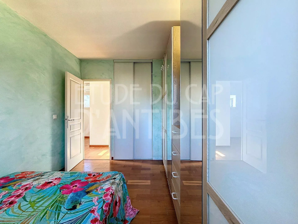 Bright bedroom with mint green walls, a colorful floral bedspread, and white wardrobes with mirrored doors along the right; an open door leads to a sunlit hallway.