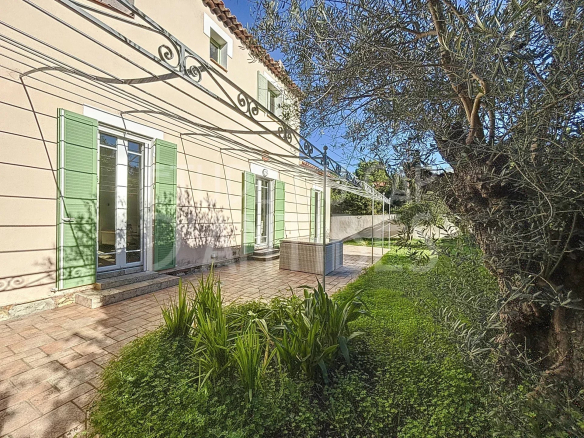 Beige house with green shutters, glass doors, and a tiled patio; olive tree on the right and garden beds in the foreground.