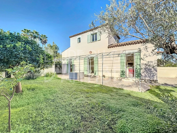 Two-story beige house with green shutters and a terracotta tile roof, set in a sunny yard with a paved patio and lush lawn. Olive trees frame the scene under a bright blue sky.