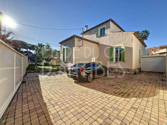 Two-story beige house with green shutters, a tiled driveway, and a black car parked near the entrance on a sunny day.