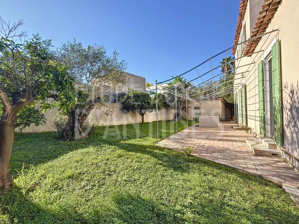 Backyard with a grassy lawn, trees, and a tiled patio along a beige house with green shutters and a metal frame for a canopy.
