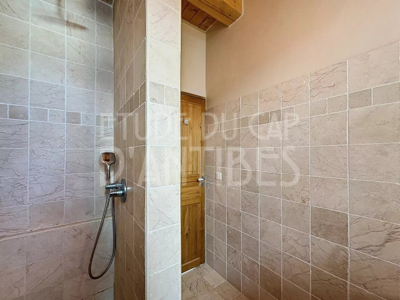 Shower area with a chrome handheld shower mounted on marble-veined tiles and a wooden door in the background.