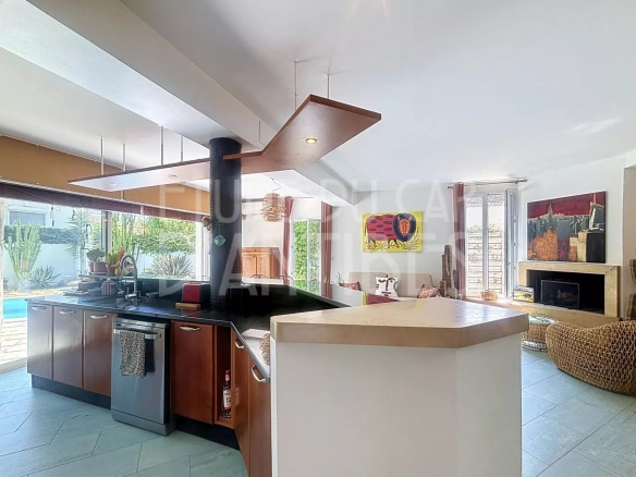 Intérieur d'une villa méditerranéenne avec cuisine ouverte et salon lumineux, piscine visible à l'extérieur.
