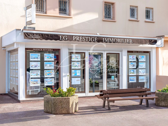 Real estate office storefront for LG Prestige Immobilier with property listings in window displays and a bench outside.