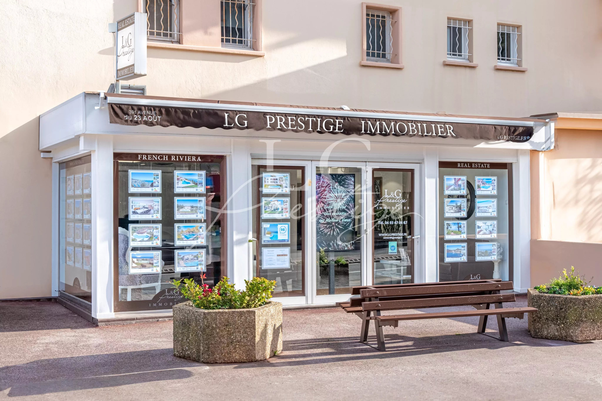 Real estate office storefront for LG Prestige Immobilier with property listings in window displays and a bench outside.