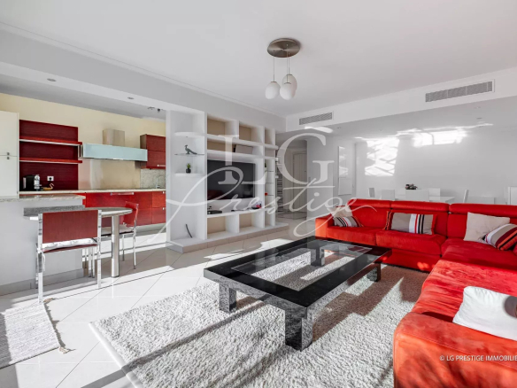Open-plan living room and kitchen with white walls, a red sectional sofa, glass coffee table, and white shelving units.