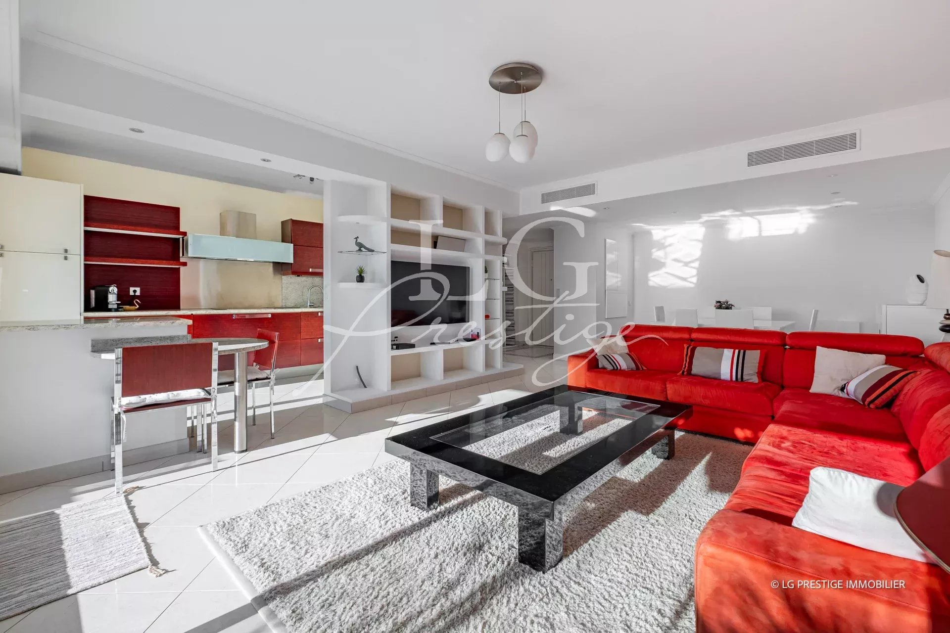 Open-plan living room and kitchen with white walls, a red sectional sofa, glass coffee table, and white shelving units.