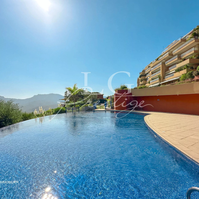 Infinity pool with curved edge and blue water, beige deck, and a hillside residential building on the right under a clear blue sky with sun glare.