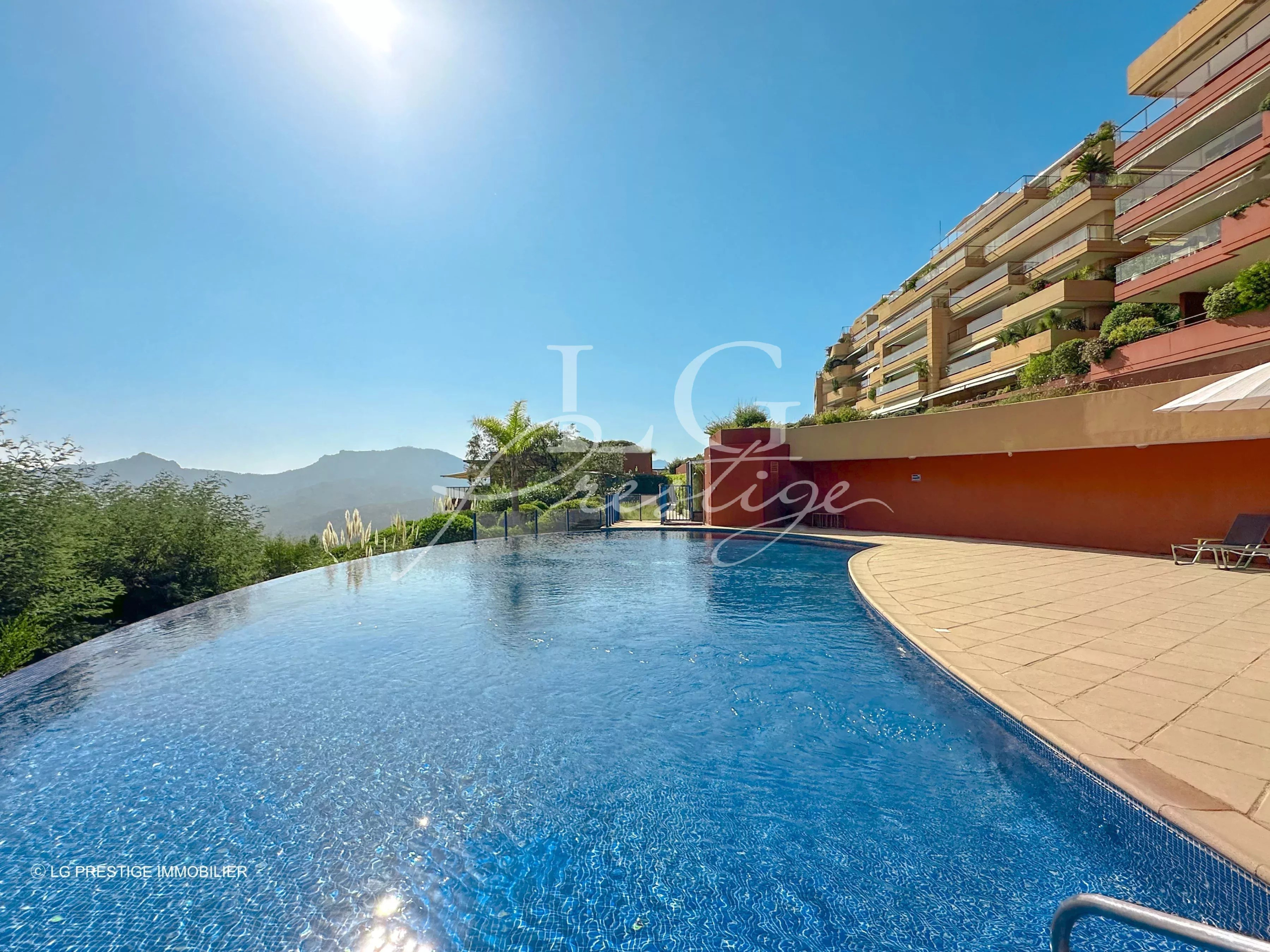Infinity pool with curved edge and blue water, beige deck, and a hillside residential building on the right under a clear blue sky with sun glare.
