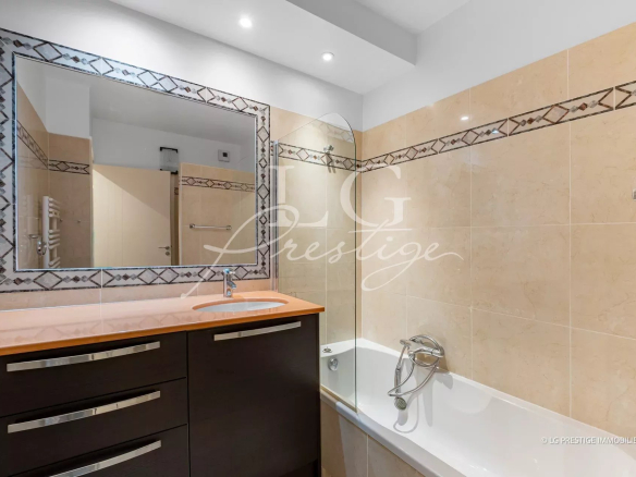 Beige tiled bathroom with a dark cabinet vanity, peach countertop, and oval sink, beside a bathtub with a curved glass panel.