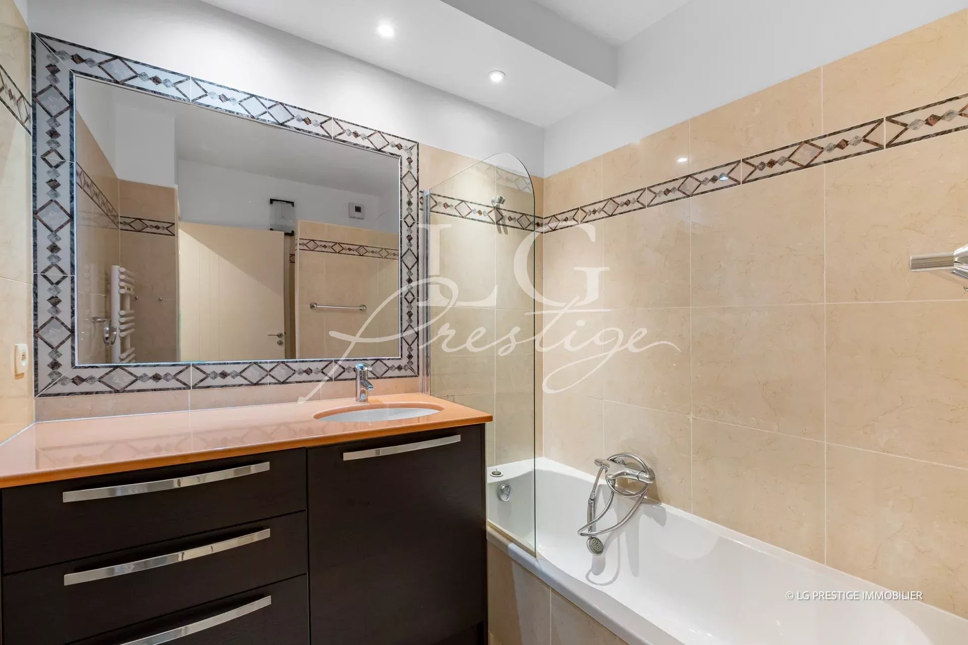 Beige tiled bathroom with a dark cabinet vanity, peach countertop, and oval sink, beside a bathtub with a curved glass panel.