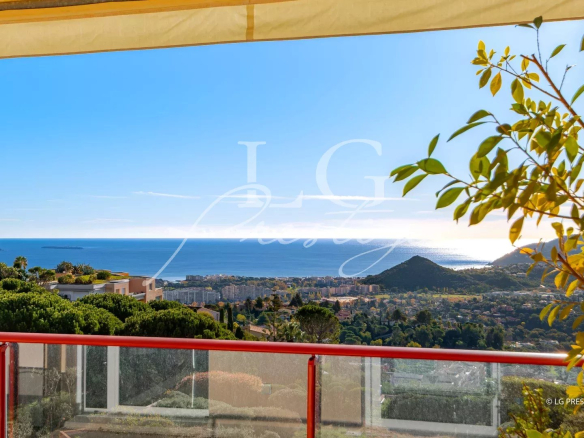 Panoramic coastal view from a balcony: blue sea, hillside houses, and green trees under a clear sky, with a red railing in the foreground and a watermark in the center.