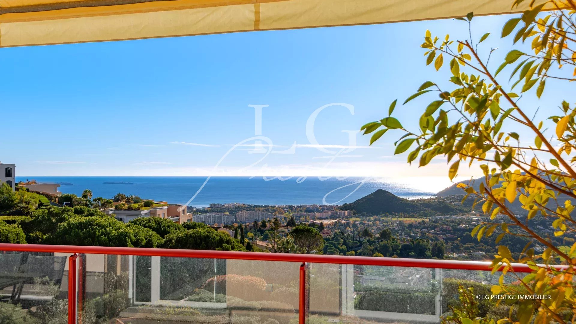 Panoramic coastal view from a balcony: blue sea, hillside houses, and green trees under a clear sky, with a red railing in the foreground and a watermark in the center.