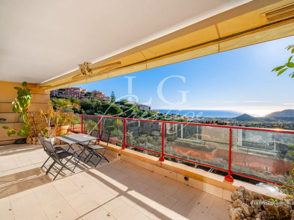 Terrace balcony with a small table and four chairs, potted plants, and a red glass railing overlooking a coastal town and ocean view.