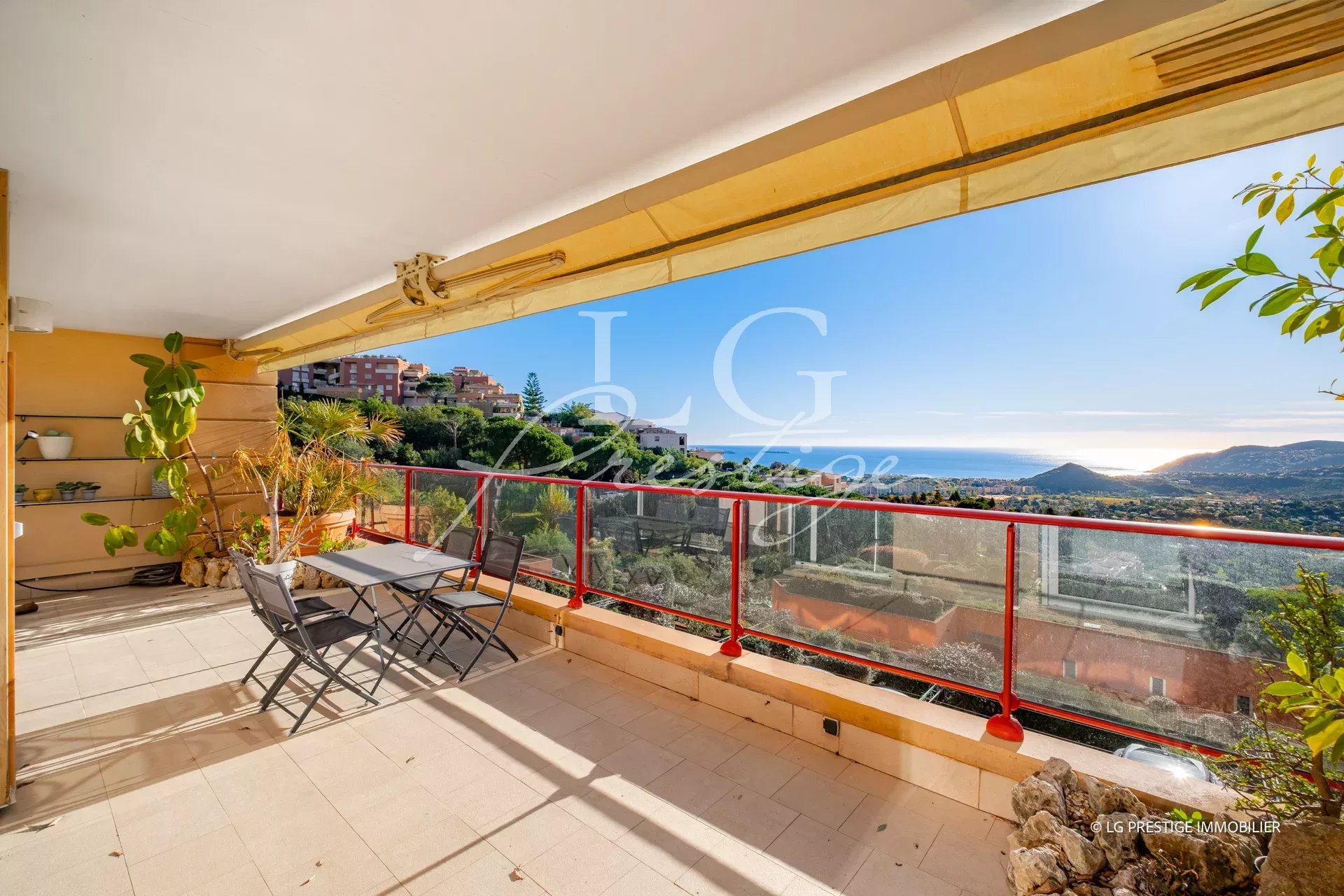 Terrace balcony with a small table and four chairs, potted plants, and a red glass railing overlooking a coastal town and ocean view.