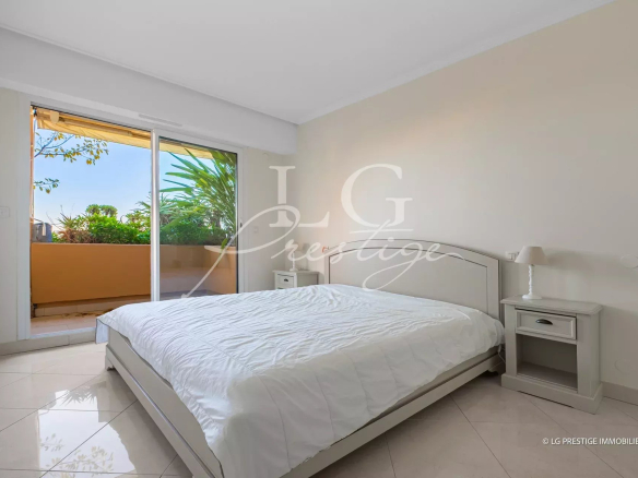 Bright bedroom with a large sliding door opening to a balcony, white bed and matching nightstands, tropical plants outside.