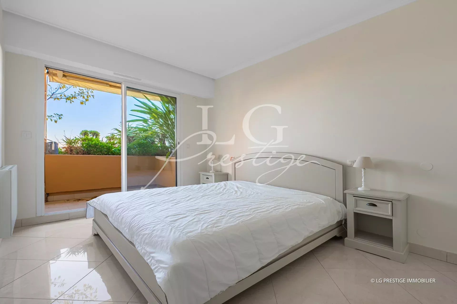 Bright bedroom with a large sliding door opening to a balcony, white bed and matching nightstands, tropical plants outside.