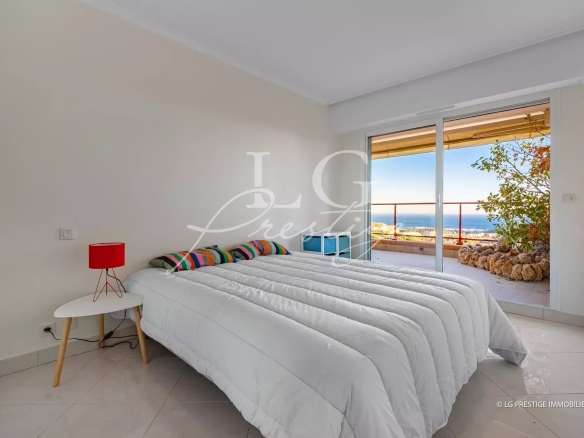 Bright bedroom with a white quilted bed, colorful striped pillows, and a red bedside lamp beside a small white table; sliding doors open to a balcony with an ocean view.