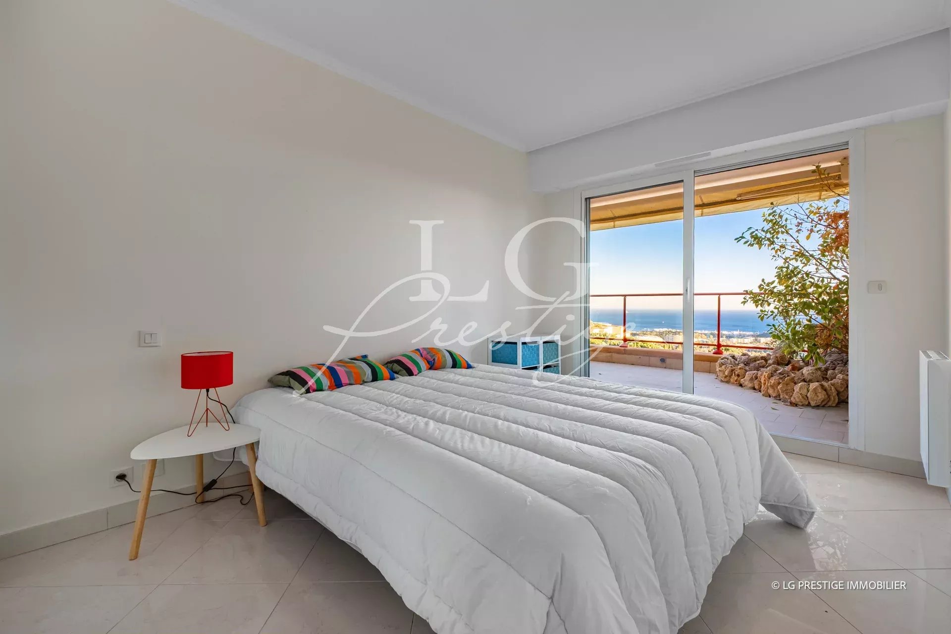 Bright bedroom with a white quilted bed, colorful striped pillows, and a red bedside lamp beside a small white table; sliding doors open to a balcony with an ocean view.
