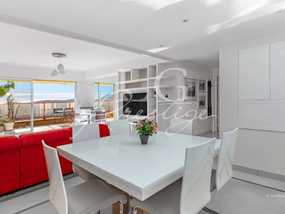 Bright open-plan living and dining area with a white dining table, white chairs, and a red sofa; sliding glass doors to a sunny balcony with potted plants.