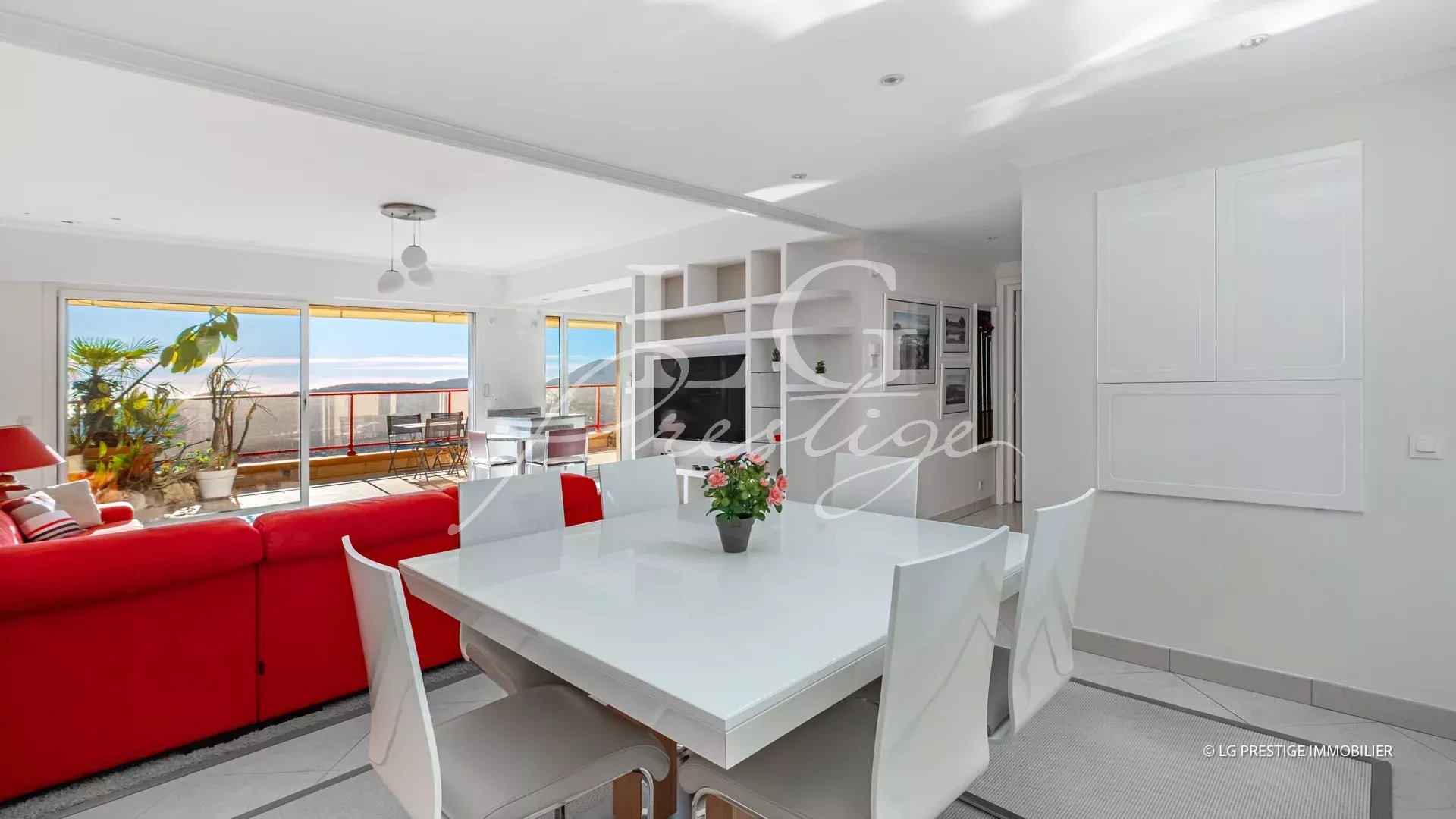 Bright open-plan living and dining area with a white dining table, white chairs, and a red sofa; sliding glass doors to a sunny balcony with potted plants.