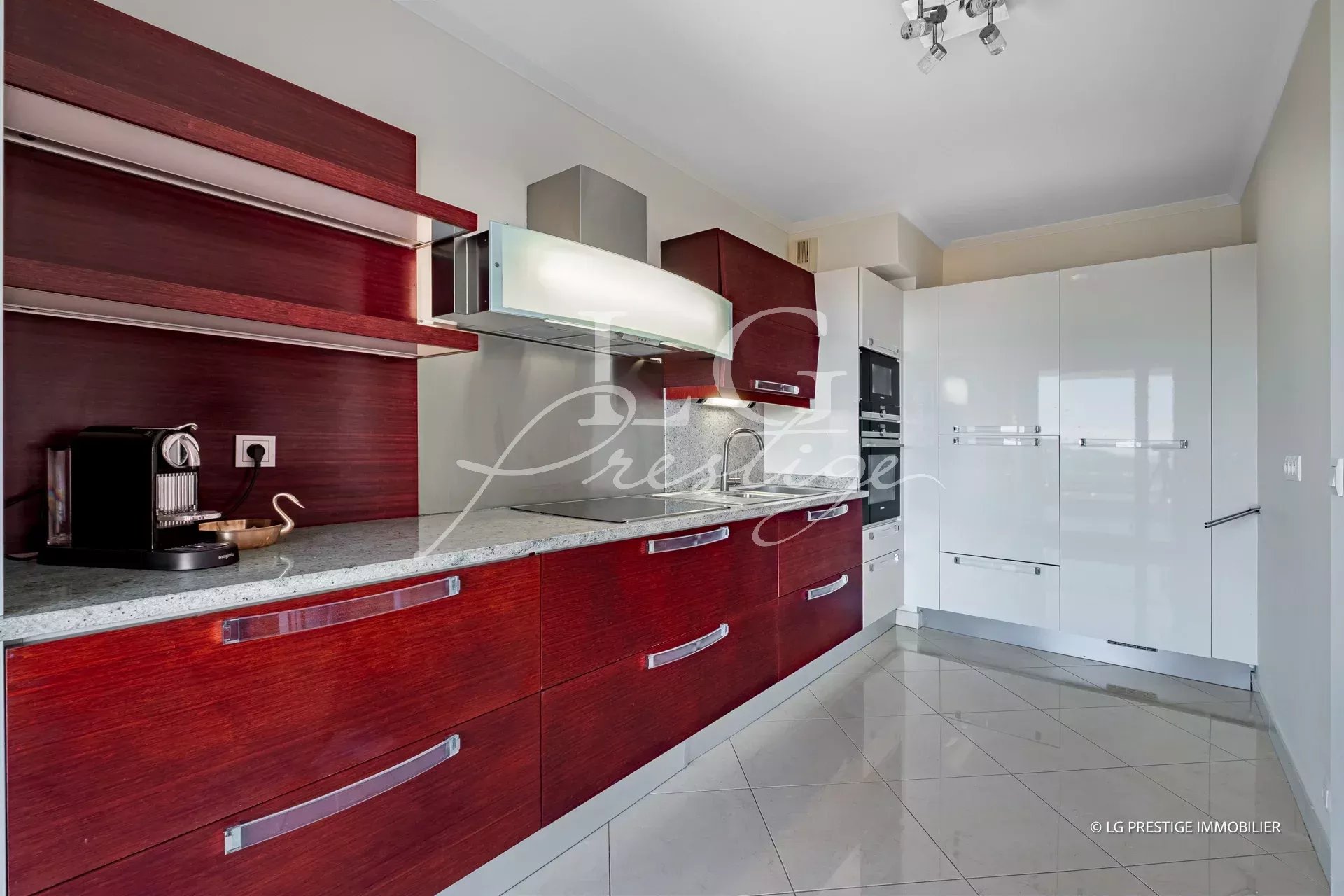 Modern kitchen with glossy white tall cabinets, dark red wood lower cabinets, and stainless steel appliances along a granite countertop.