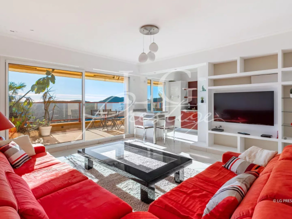 Bright living room with a red sectional sofa, glass coffee table, and built-in white shelving with a wall TV, open to a sunny balcony with ocean view.