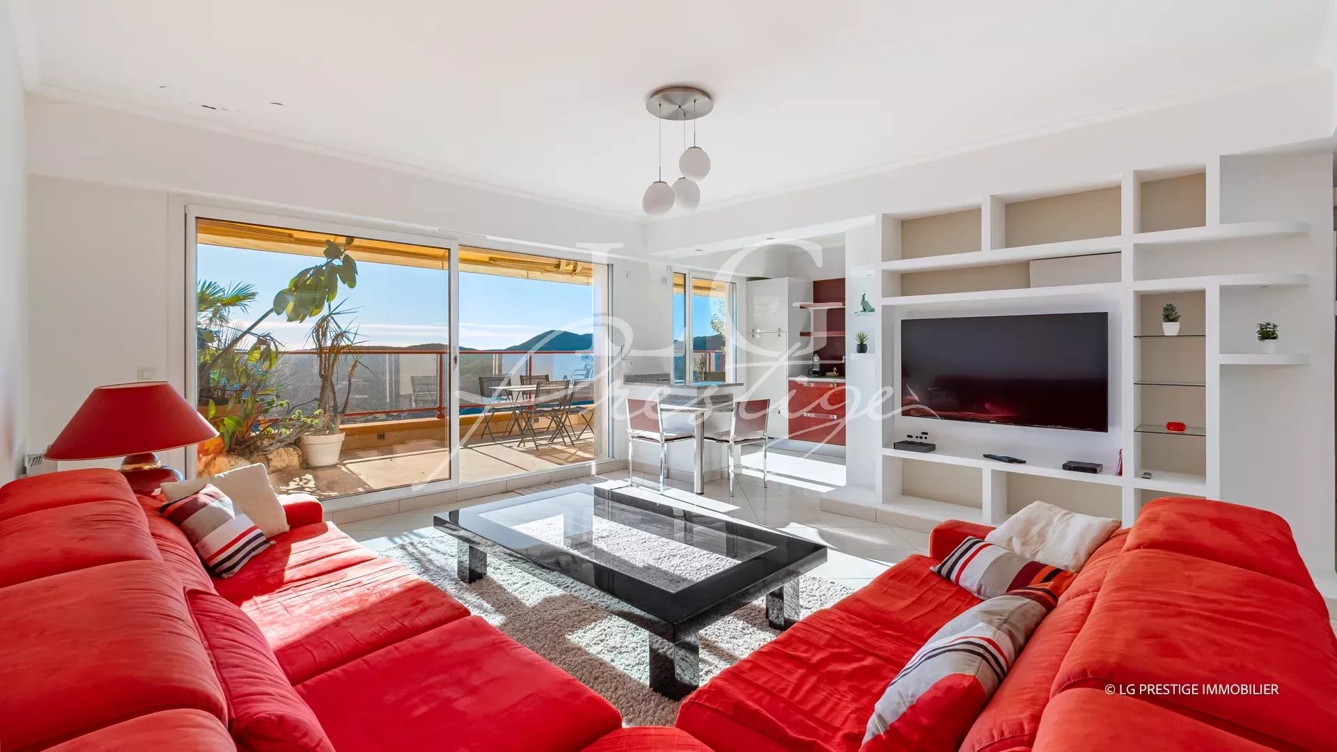 Bright living room with a red sectional sofa, glass coffee table, and built-in white shelving with a wall TV, open to a sunny balcony with ocean view.
