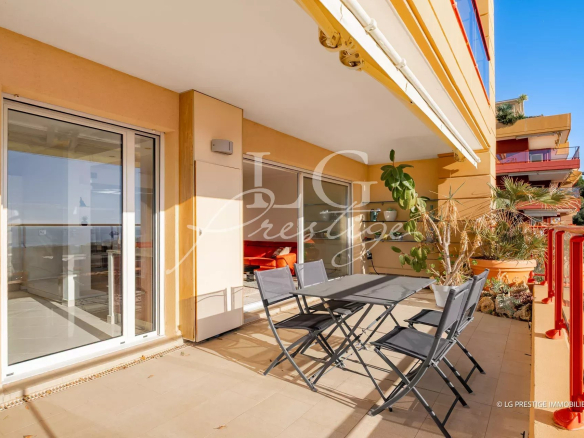 Outdoor apartment balcony with a gray dining table and six folding chairs, potted plants, and sliding glass doors to the interior.