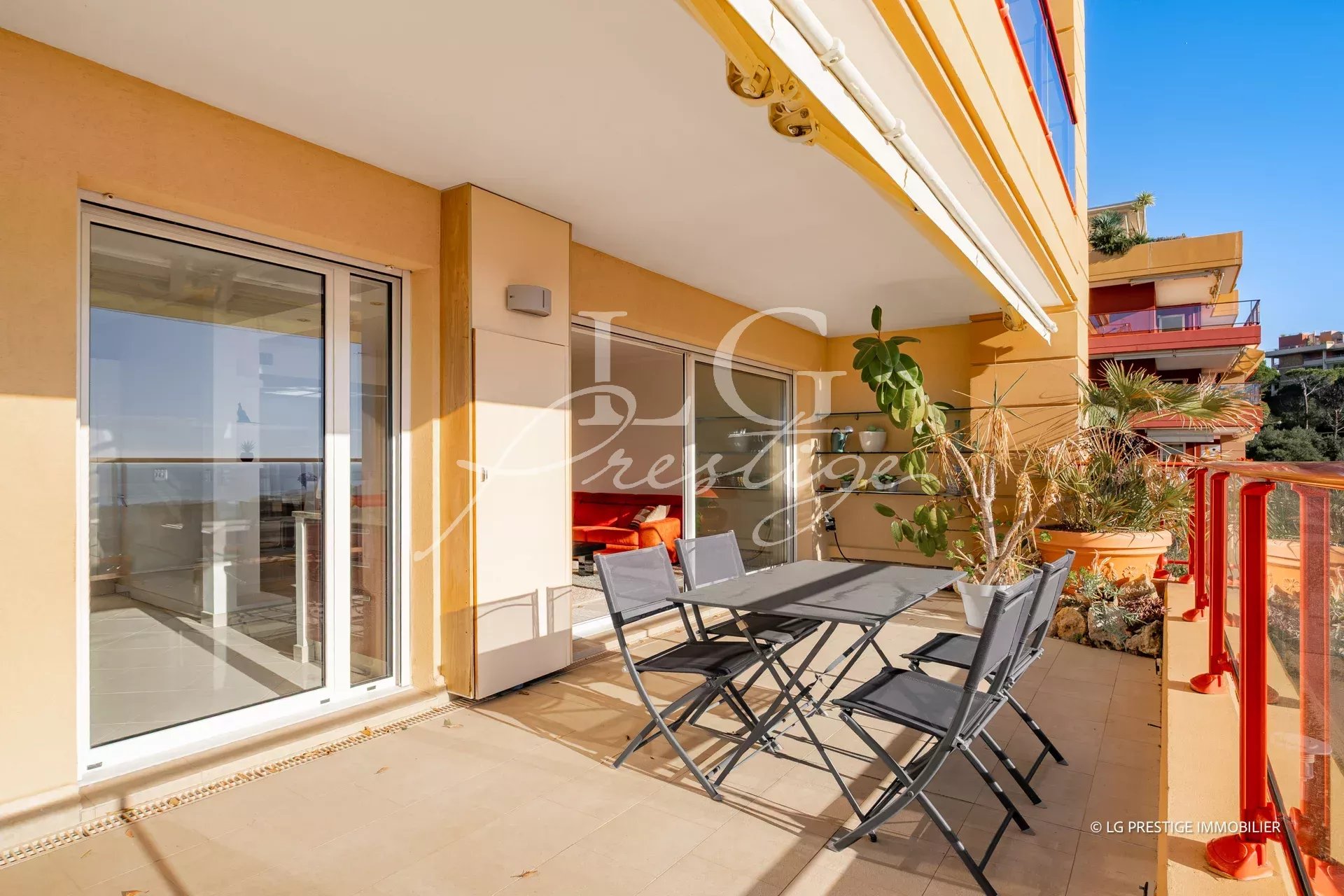 Outdoor apartment balcony with a gray dining table and six folding chairs, potted plants, and sliding glass doors to the interior.
