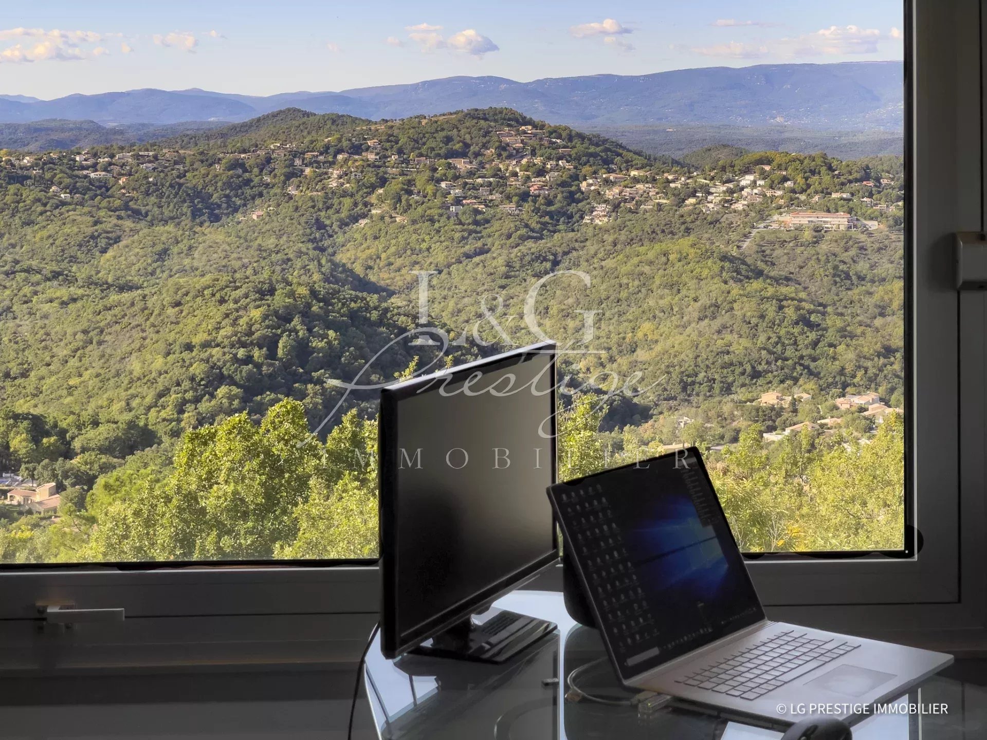 Vue panoramique sur les collines verdoyantes depuis une fenêtre avec un bureau et des ordinateurs.