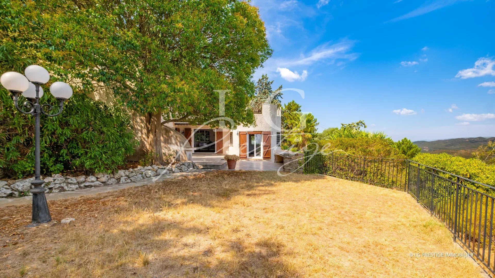 View of a villa with garden and trees in Théoule-sur-Mer, blue sky and clouds.