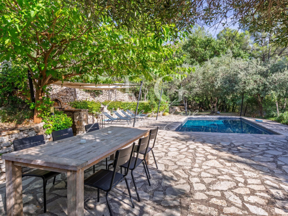 Stone terrace with wooden table, chairs, swimming pool and lush vegetation around.