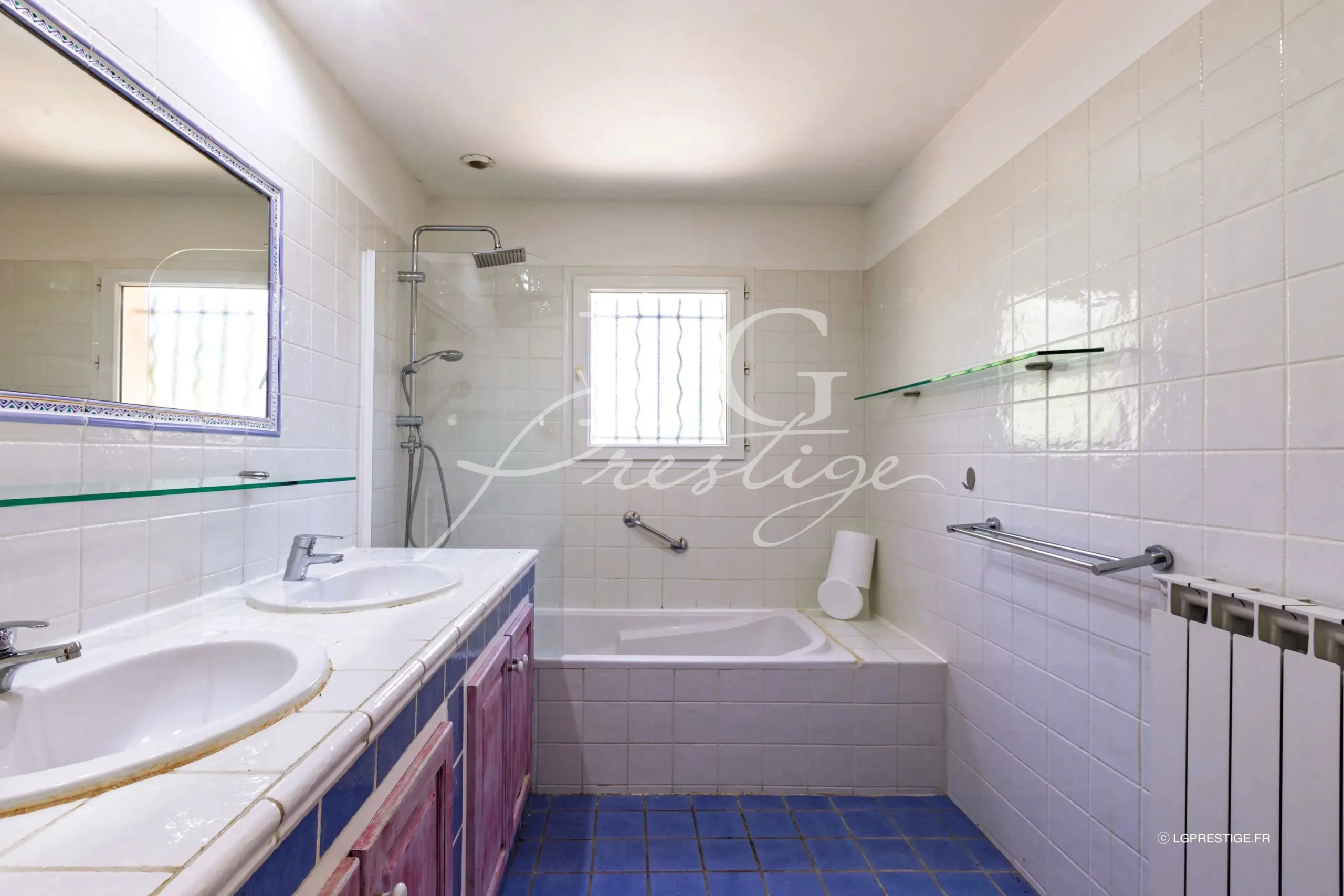 Salle de bain avec baignoire, lavabo double et douche, carrelage blanc et bleu.