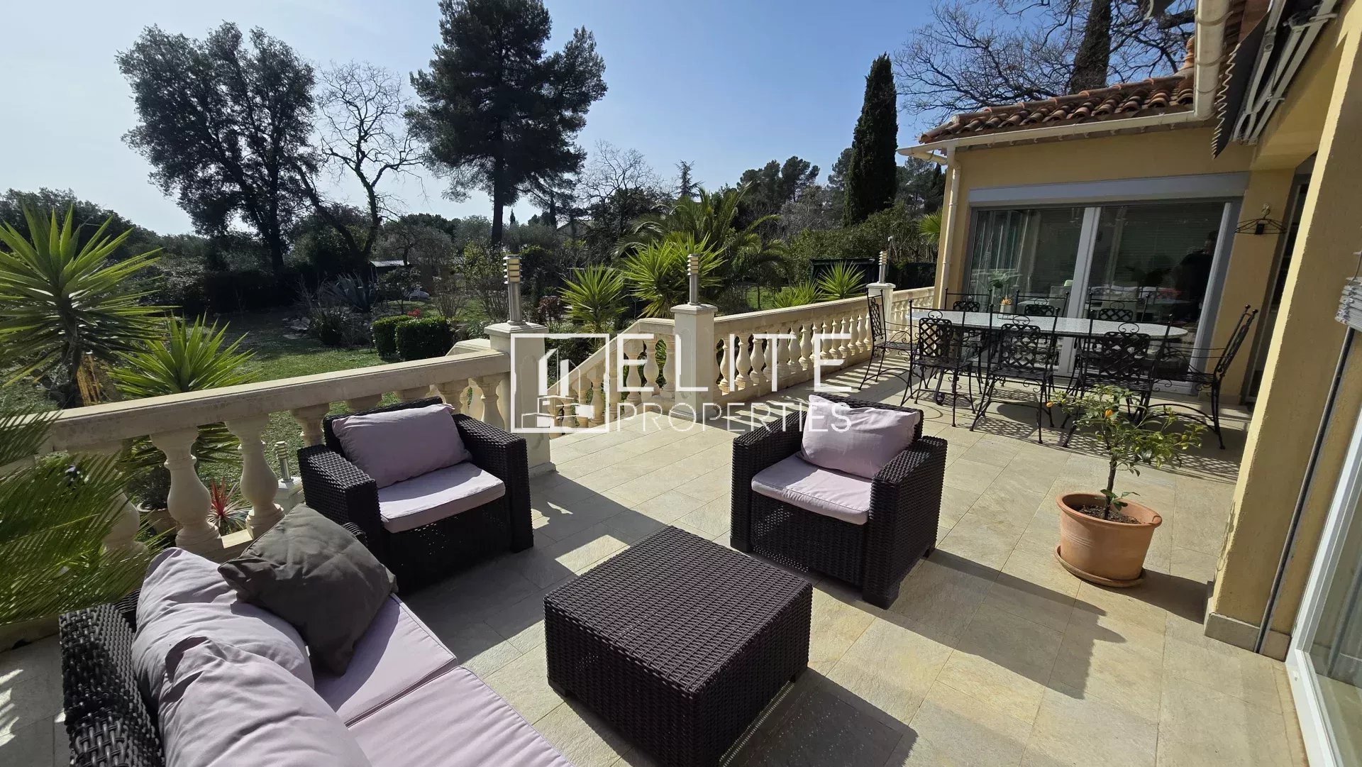 Outdoor terrace with wicker seating area and beige cushions overlooking a lush garden. A glass door leads to the interior.
