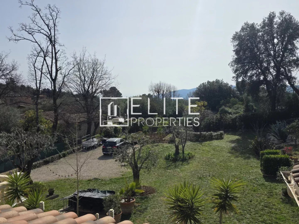Overhead view of a spacious backyard with trees, a grassy lawn, and a gravel driveway; a dark SUV is parked near the center, with tiled roof and patio area in the foreground and a hillside beyond. (Elite Properties watermark)