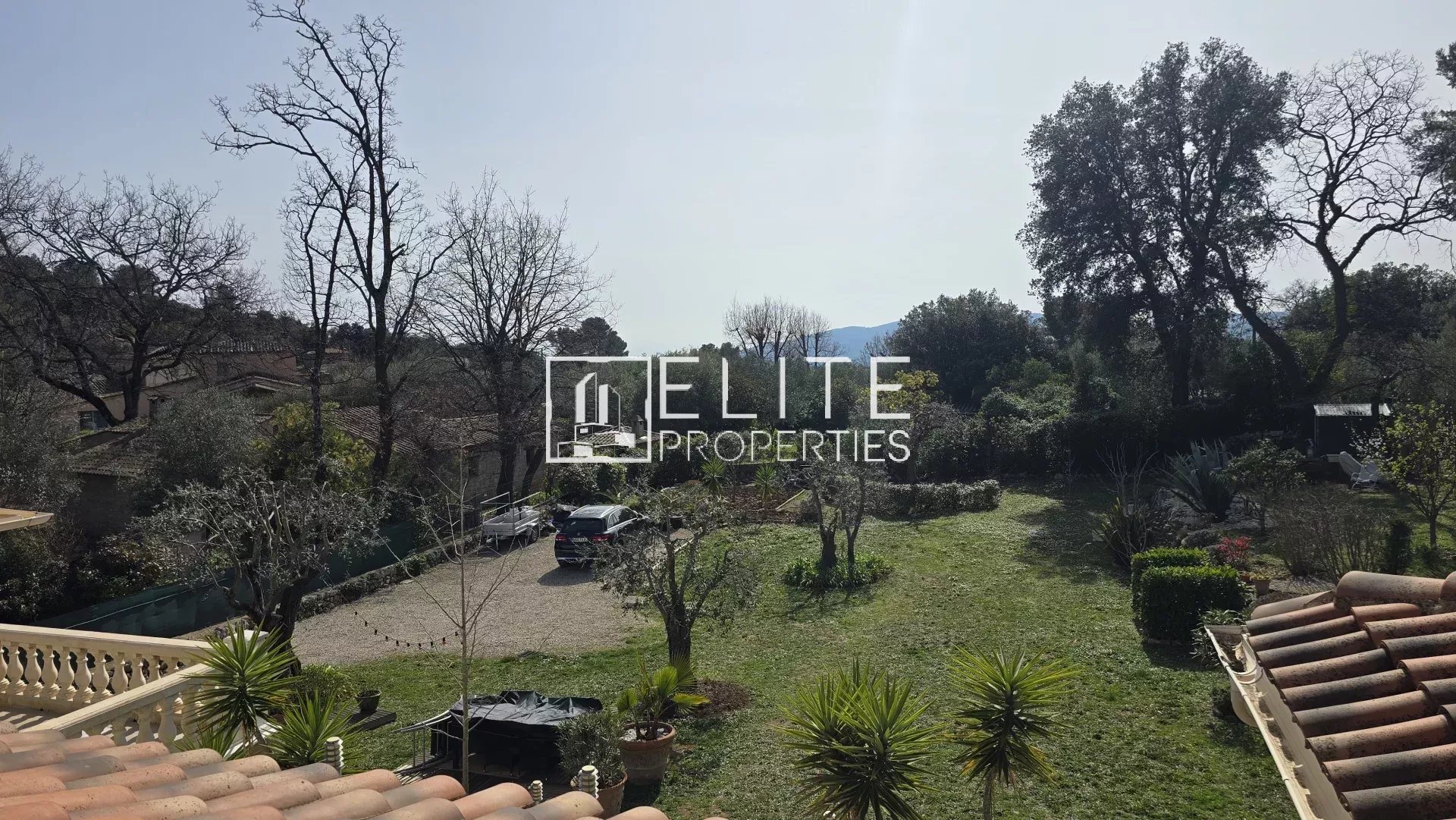 Overhead view of a spacious backyard with trees, a grassy lawn, and a gravel driveway; a dark SUV is parked near the center, with tiled roof and patio area in the foreground and a hillside beyond. (Elite Properties watermark)