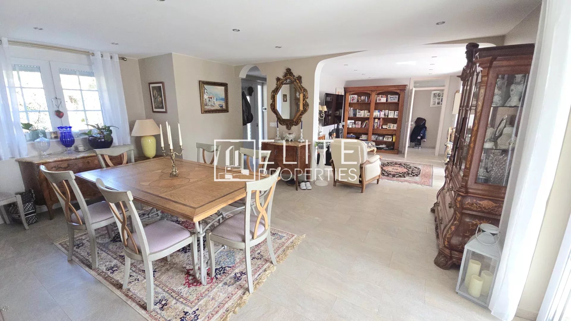 Spacious dining area with a wooden table and six chairs, beige walls, and decorative antiques along a sideboard and mirror.