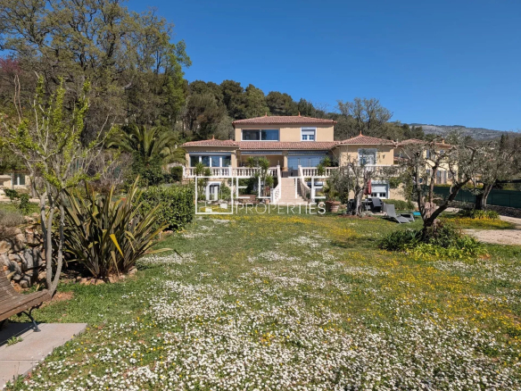 Two-story Mediterranean villa with beige walls, terracotta roof, and balconies, set in a garden with white and yellow flowers under a blue sky.