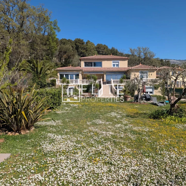 Two-story Mediterranean villa with beige walls, terracotta roof, and balconies, set in a garden with white and yellow flowers under a blue sky.