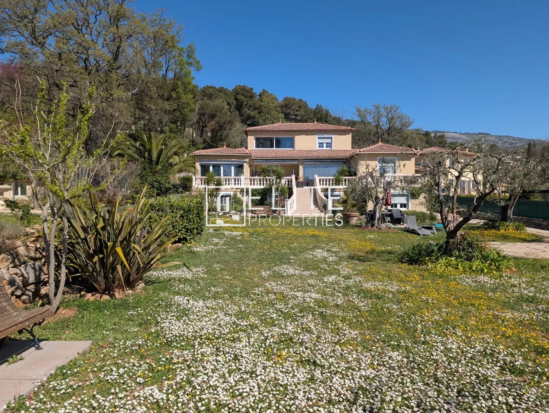 Two-story Mediterranean villa with beige walls, terracotta roof, and balconies, set in a garden with white and yellow flowers under a blue sky.
