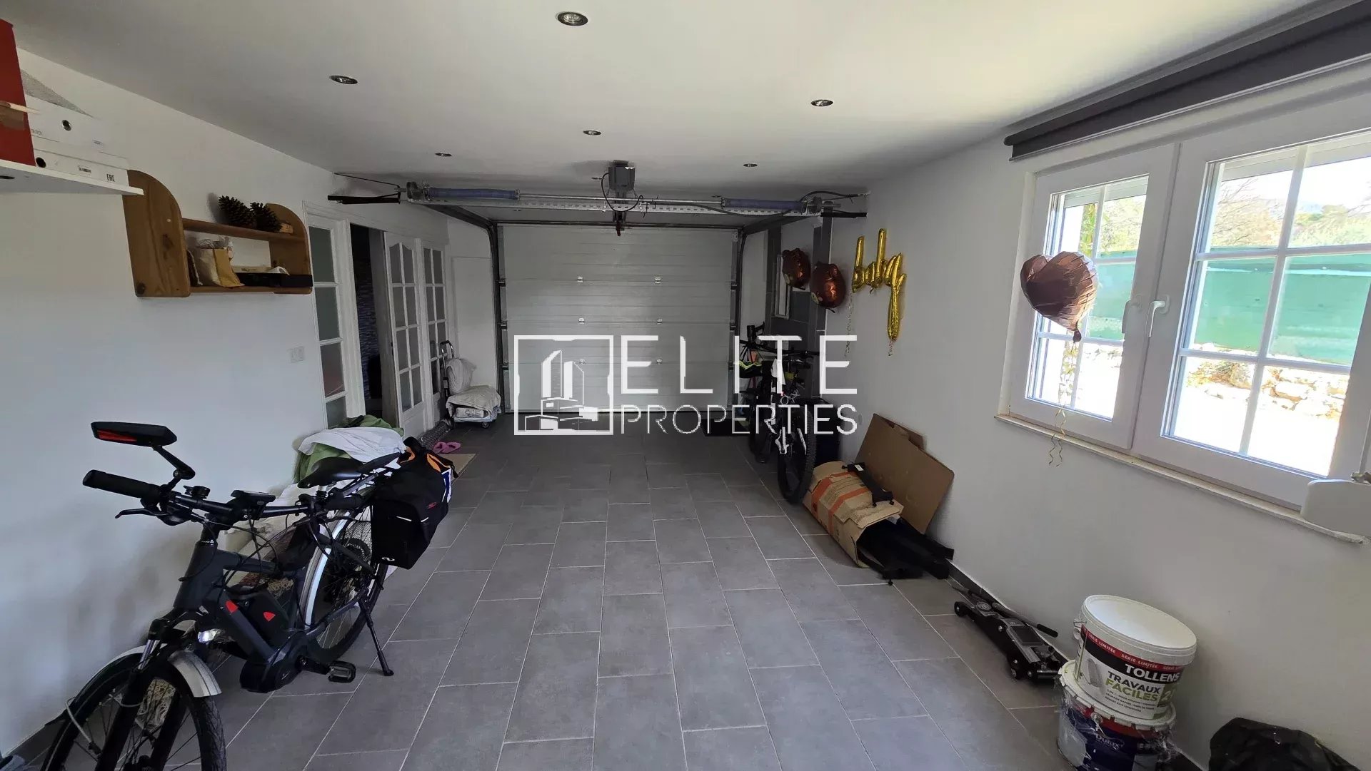 Bright home garage with bicycles on the left, storage shelves along the wall, a white sectional door at the back, and a window with gold '4th' balloons on the right.